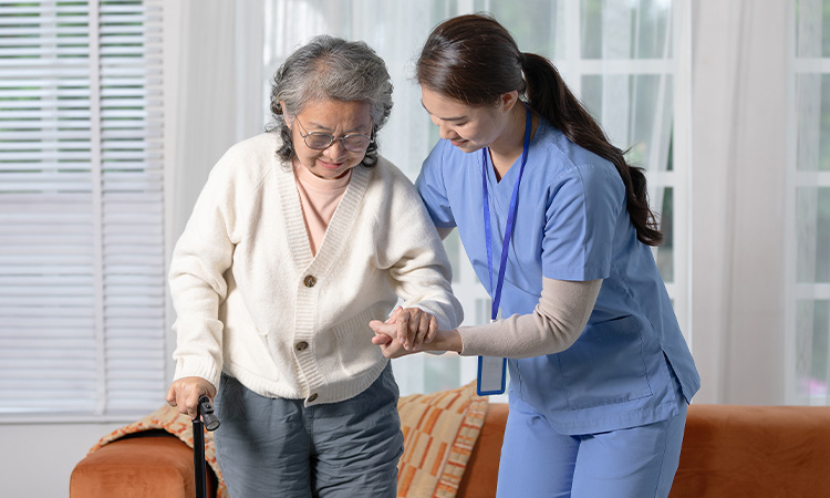 Caregiver in blue scrubs assisting an elderly lady walking with a walking stick