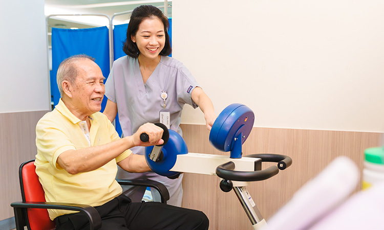 An elderly man in a yellow shirt uses an arm exercise machine, assisted by a smiling nurse in scrubs.