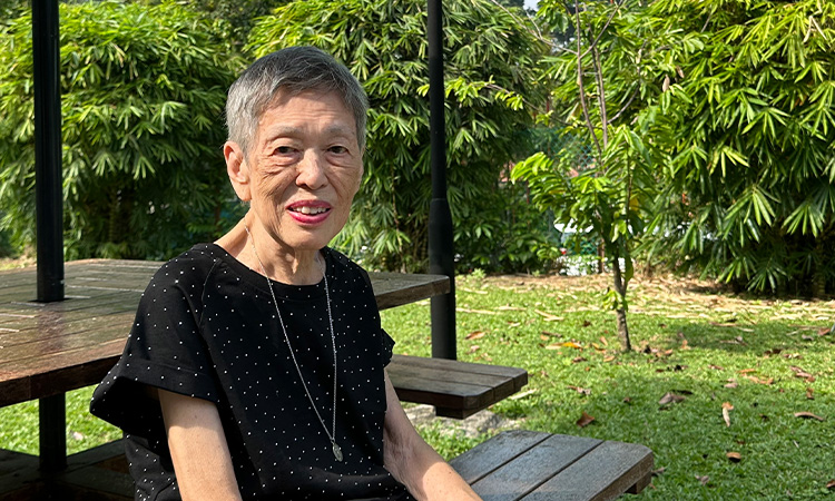 An elderly woman with short gray hair, wearing a black dress, sits on a wooden bench.