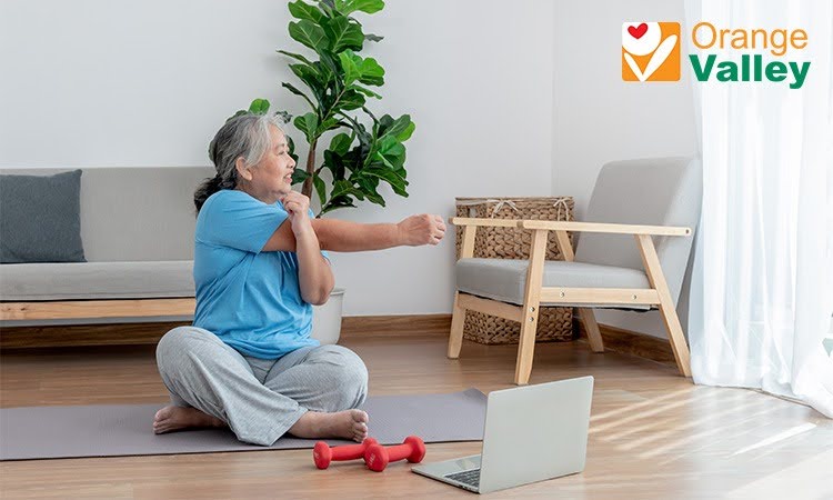 An elderly Asian woman stretching her arm across her chest while sitting on a yoga mat at home.