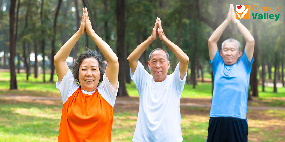 Three smiling Asian seniors doing a yoga pose outdoors in a park.
